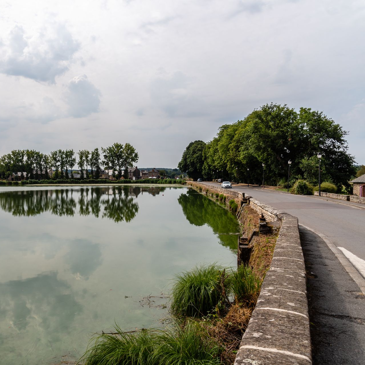 Un chemin de pierre sinueux longe un lac calme, avec des arbres, un ciel nuageux et des reflets sur l'eau.