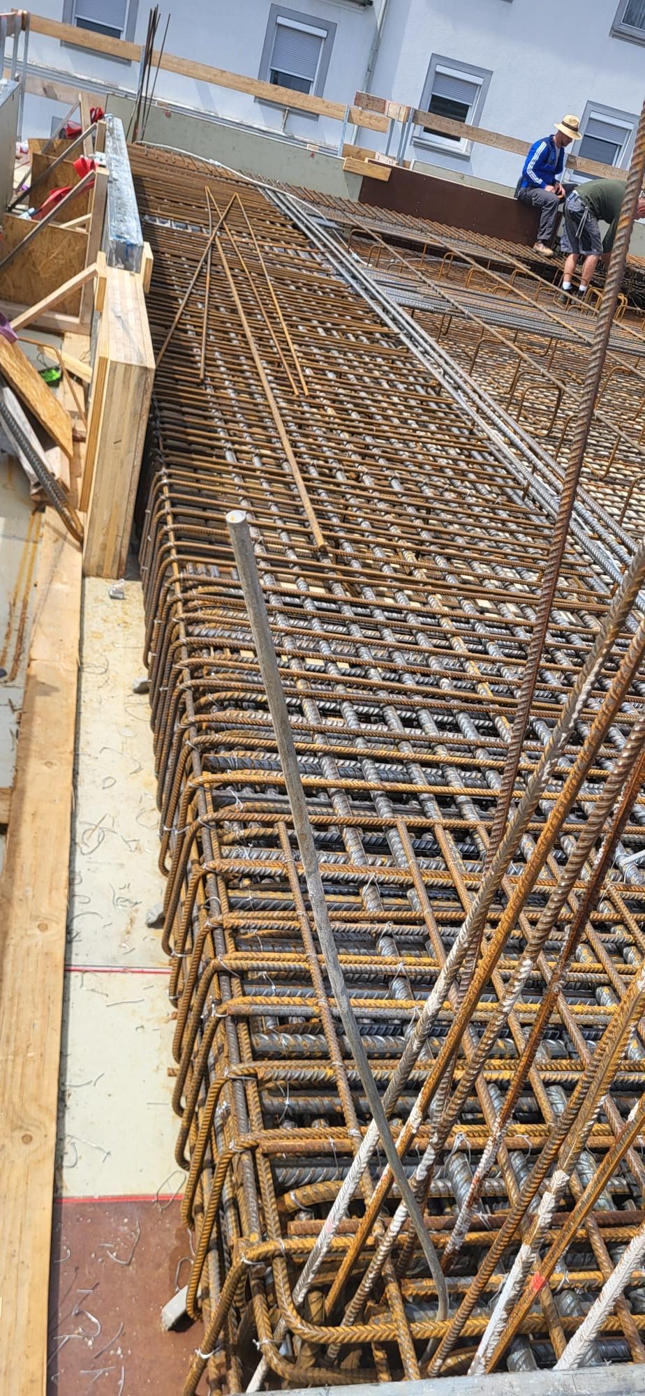 Baustelle mit Stahlbewehrungsgitter für eine Betonplatte neben einem Gebäude; im Hintergrund sind Arbeiter zu sehen.