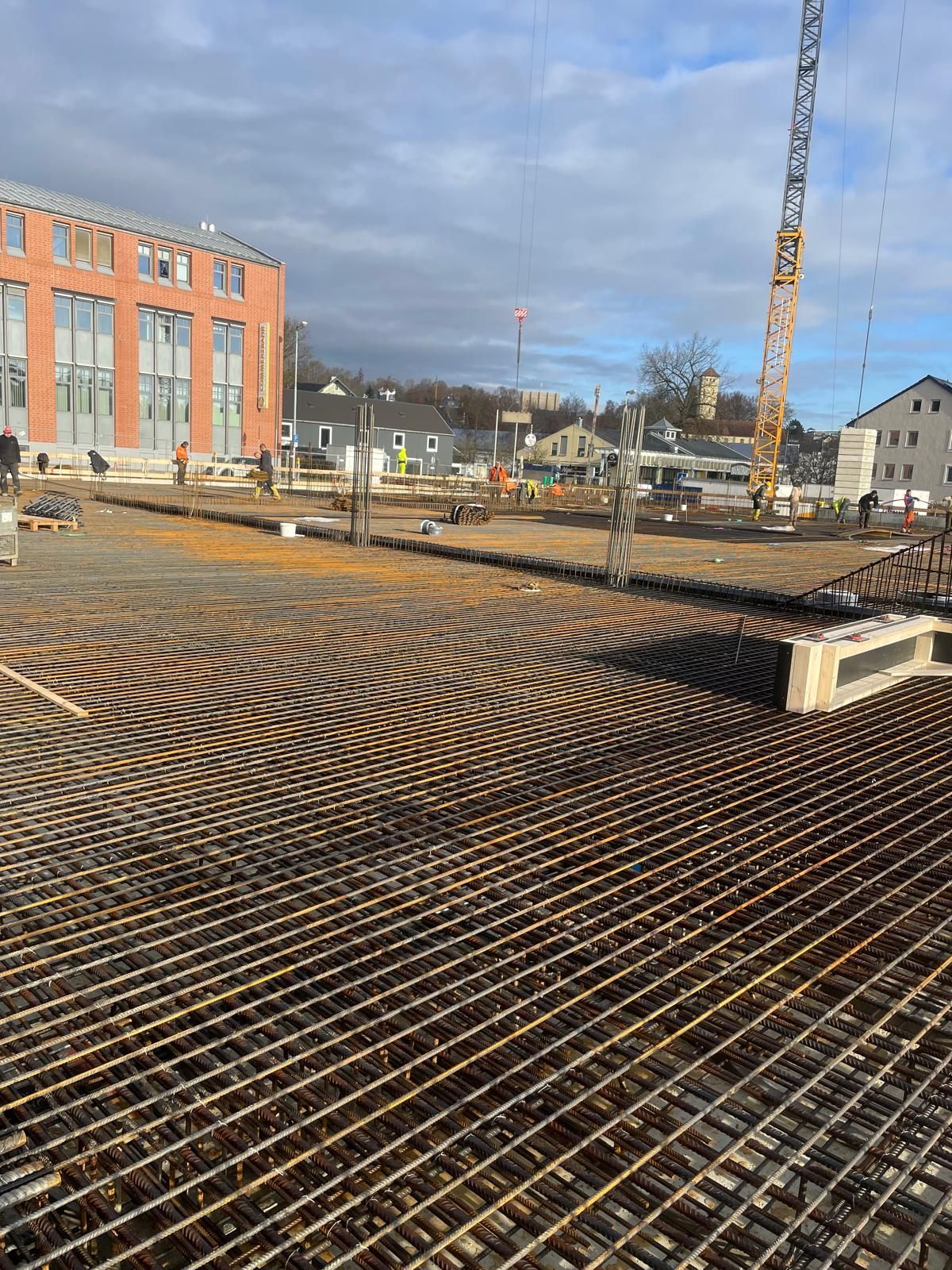 Baustelle mit Bewehrungsgittern, Kran und teilweise errichteten Gebäuden unter bewölktem Himmel.