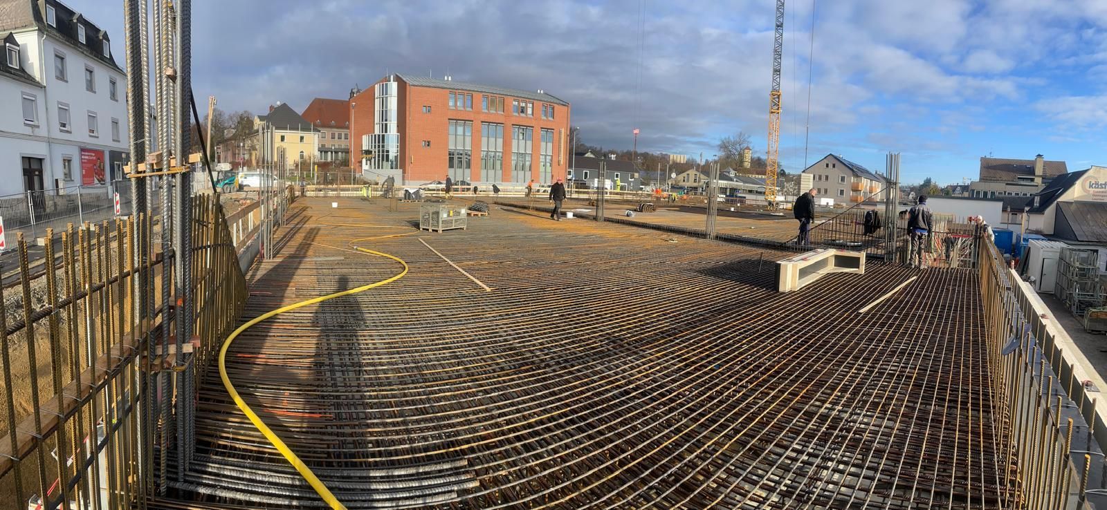 Baustelle mit freiliegendem Bewehrungsstahl, Gebäude im Hintergrund, gelber Schlauch, Kran, bewölkter Himmel.