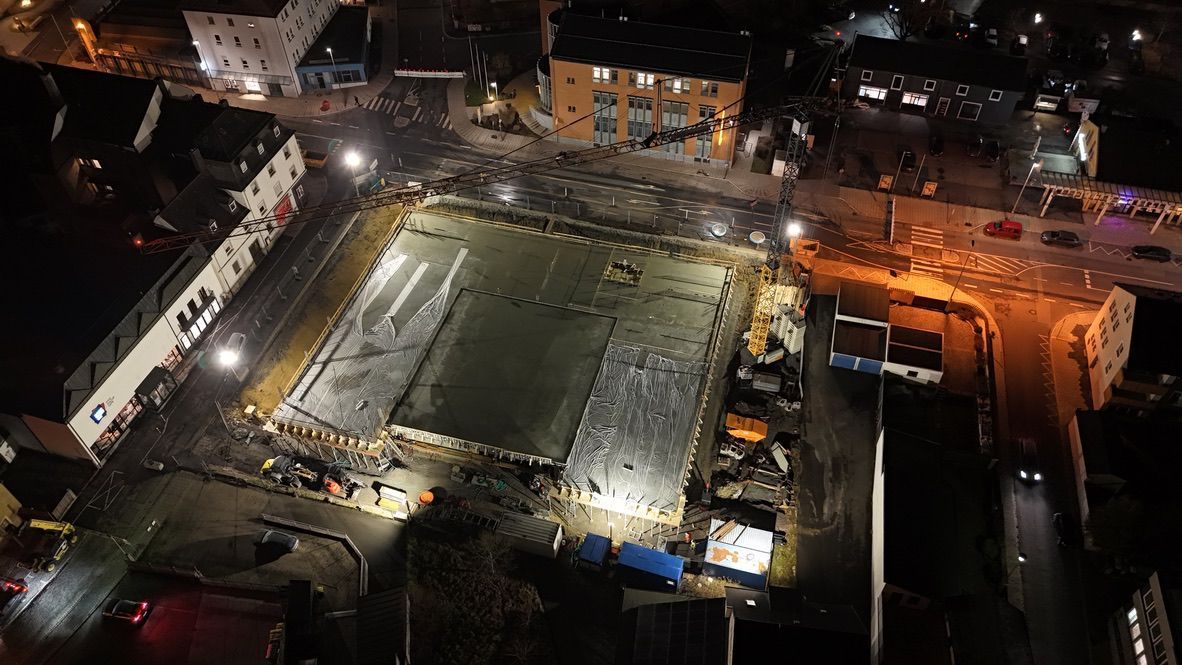 Luftaufnahme einer Baustelle bei Nacht mit umliegenden Gebäuden. Flutlichtstrahler beleuchten das Gelände.
