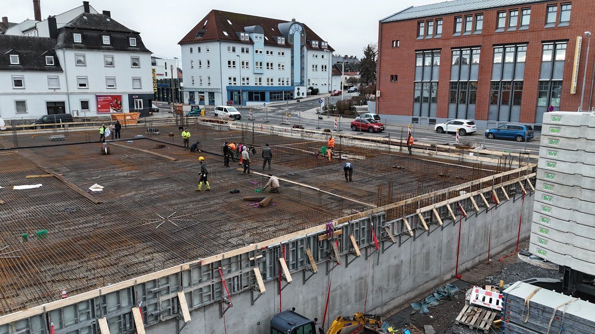 Baustelle mit Arbeitern, Bewehrungsstahl, Betonschalungen und Gebäuden im Hintergrund.