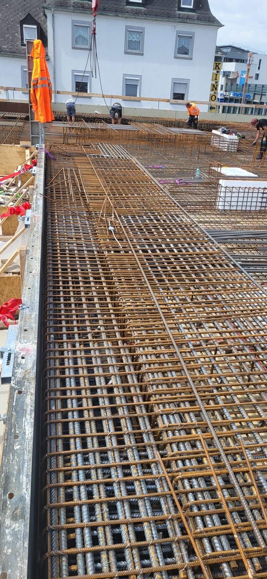Baustelle mit Stahlbewehrungsgitter über einem Fundament, neben einem weißen Haus und einem Arbeiter in orangefarbener Weste.