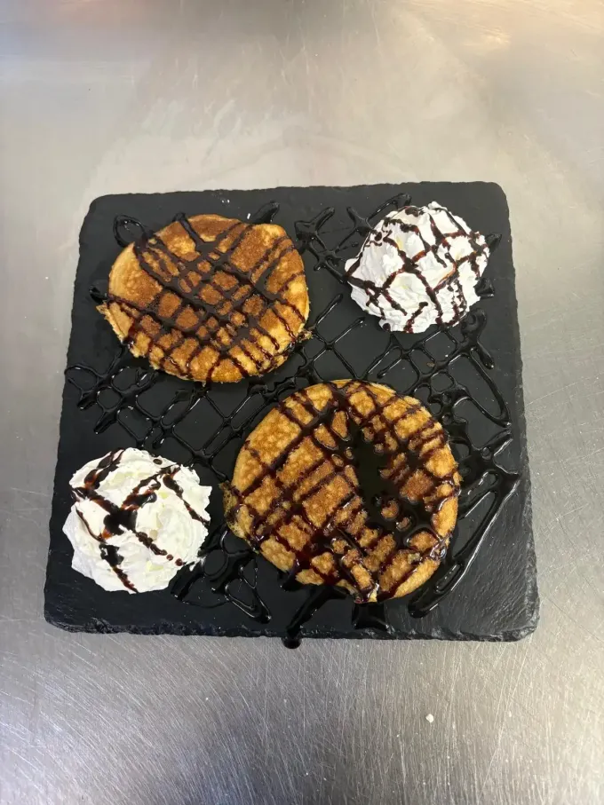 Dos panqueques bañados en jarabe y coronados con cucharadas de crema batida sobre una tabla de pizarra oscura.