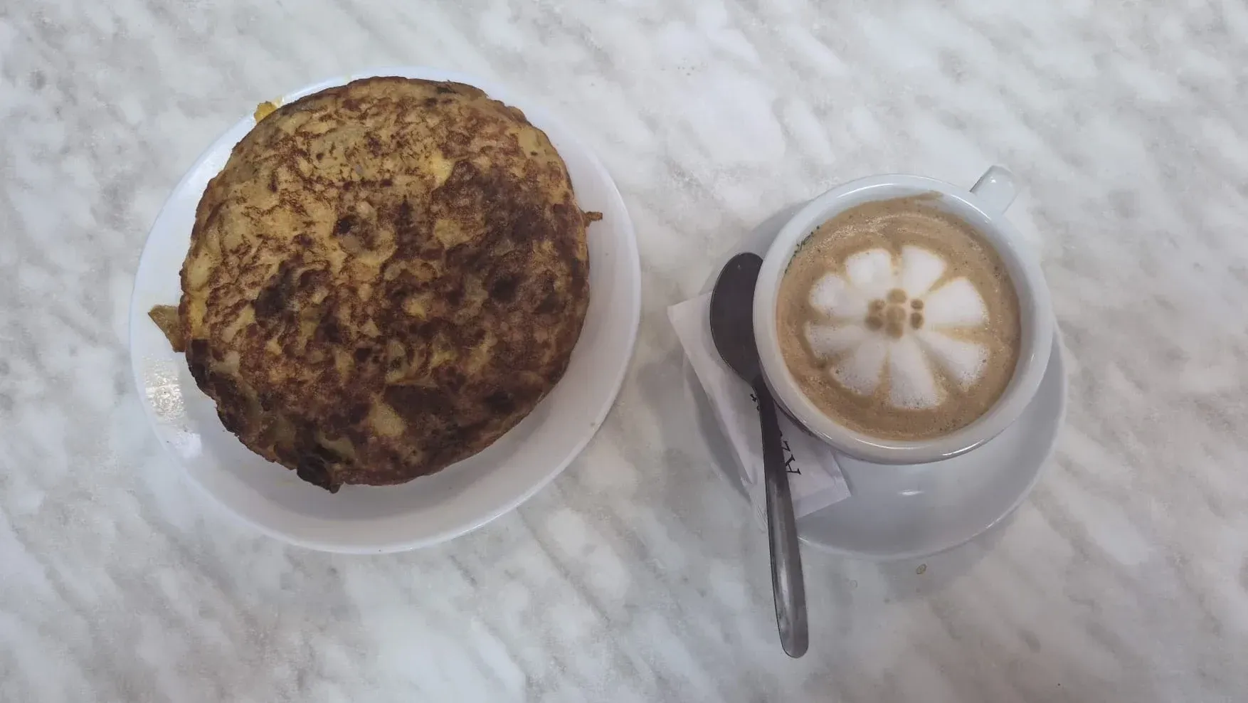 Una taza de café con arte latte junto a una magdalena sobre una mesa blanca con una cuchara.