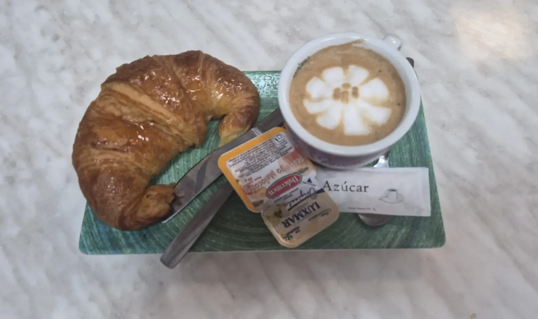 Croissant y café con leche en una bandeja con sobres de azúcar y una cuchara, vistos desde arriba.