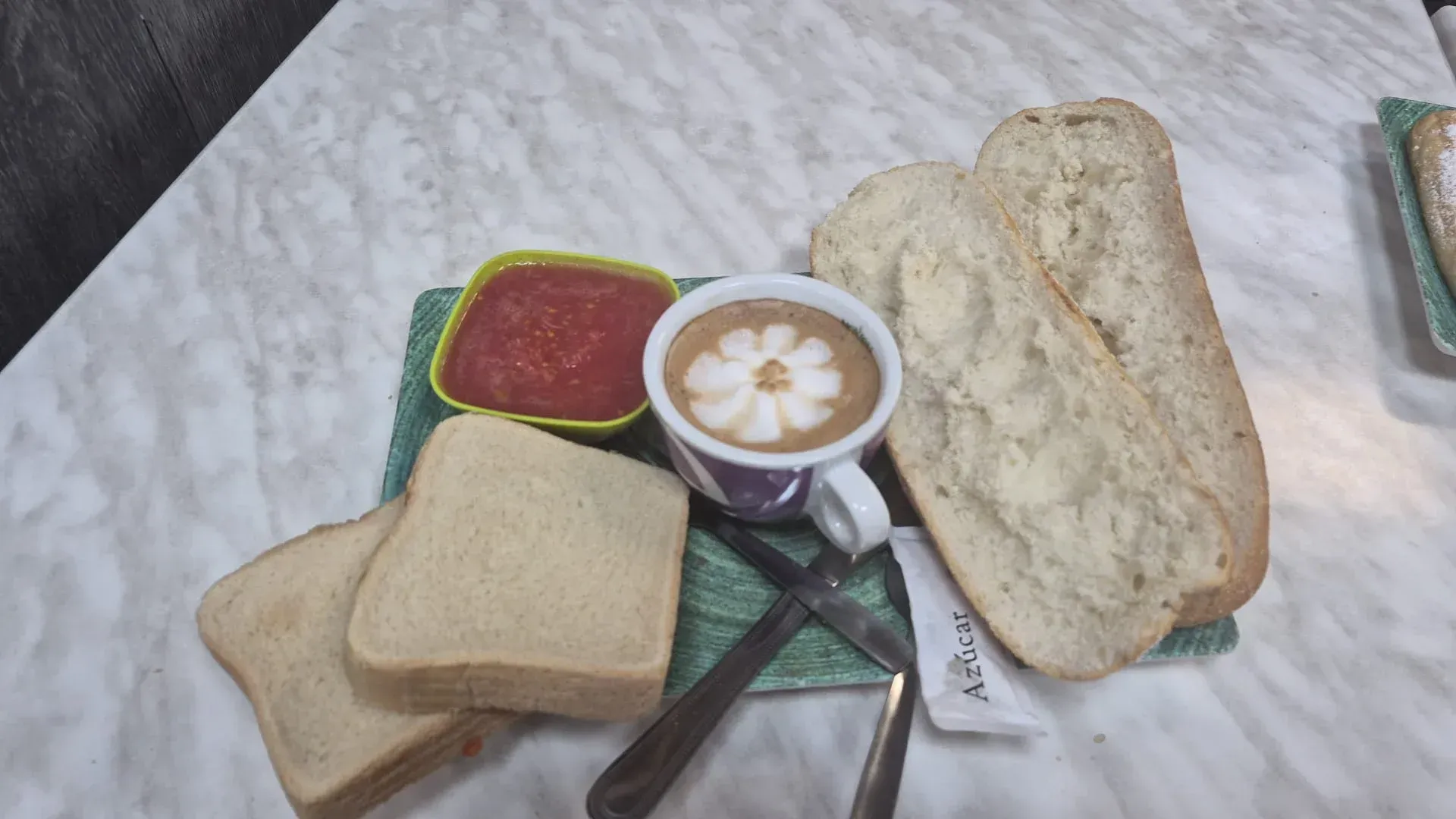 Variedad de panes, un café con leche y una salsa roja en una bandeja con cubiertos sobre una mesa.
