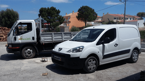 Uma carrinha Peugeot branca e um pequeno camião estacionados ao ar livre. Edifícios e árvores ao fundo.