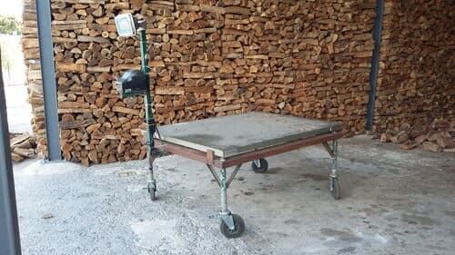 Uma mesa de trabalho com rodinhas e luminária, em frente a uma pilha de lenha. Dentro de um prédio com vigas de metal.