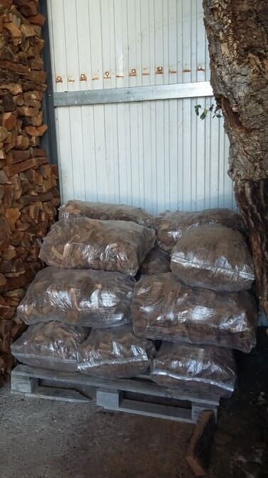 Sacos de material marrom sobre um palete dentro de um prédio. Pilha de lenha à esquerda.