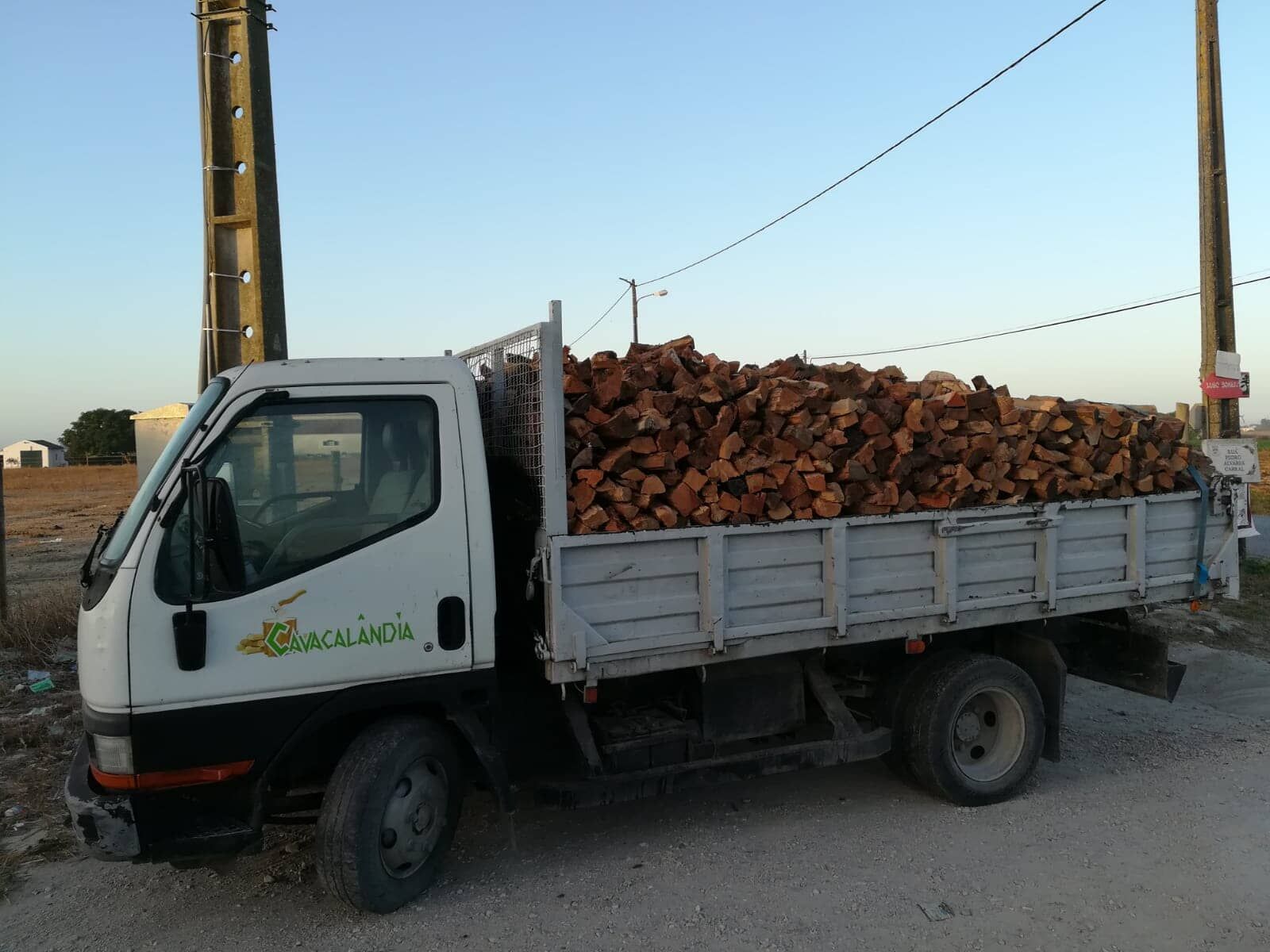 Carroceria de caminhonete cheia de lenha, estacionada em uma estrada de cascalho, sob um céu azul.