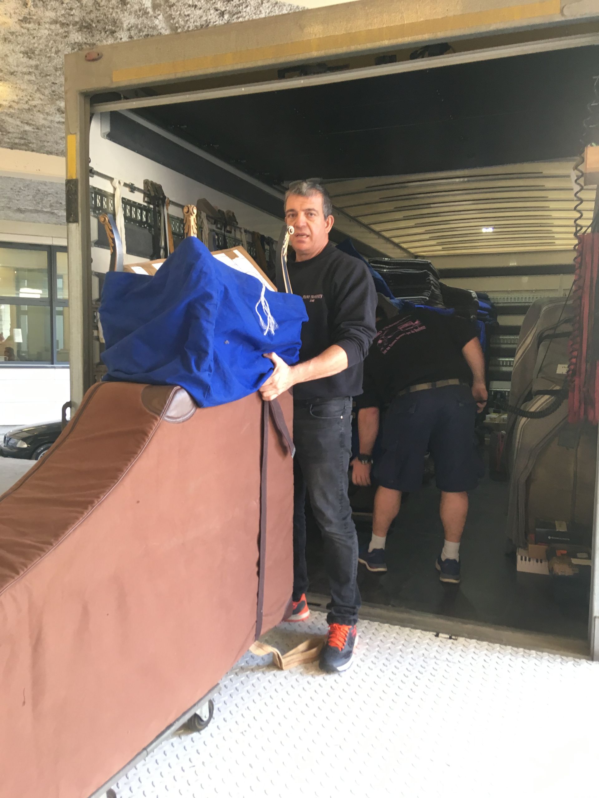 Un homme pousse un objet recouvert d'un film bleu sur un chariot, le déchargeant d'un camion ou d'une remorque et le faisant entrer dans un garage.
