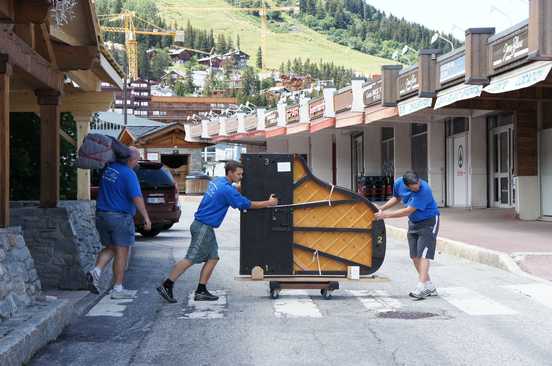 Trois personnes en chemises bleues déplacent un piano sur un chariot dans une rue d'un village de montagne.