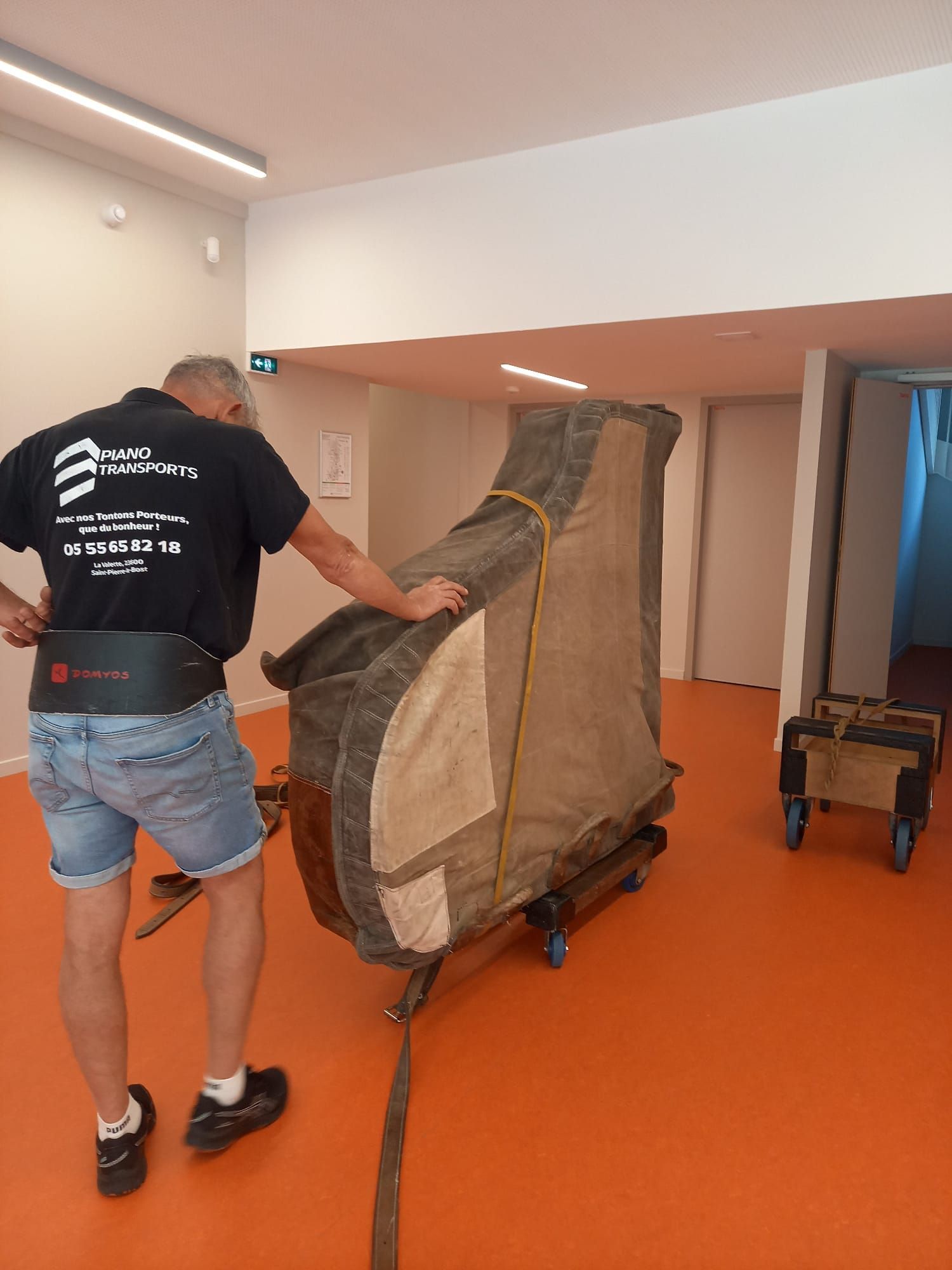Un ouvrier pousse un canapé emballé sur un chariot dans un couloir au sol orange.