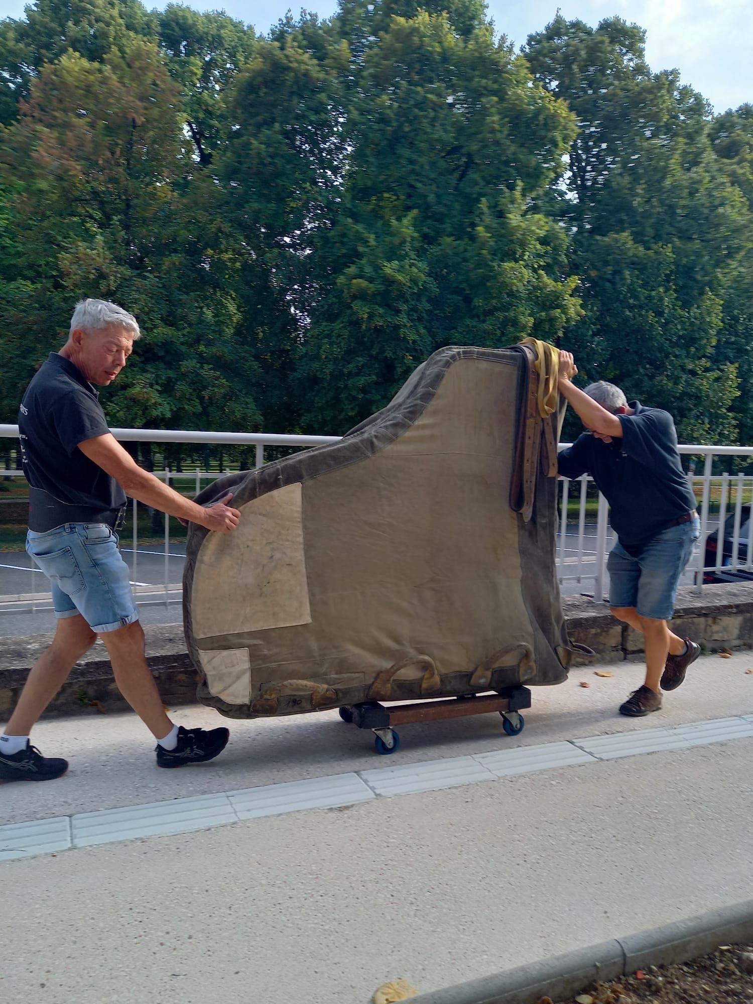 Deux personnes poussent un grand canapé sur un trottoir, avec des arbres et un pont en arrière-plan.