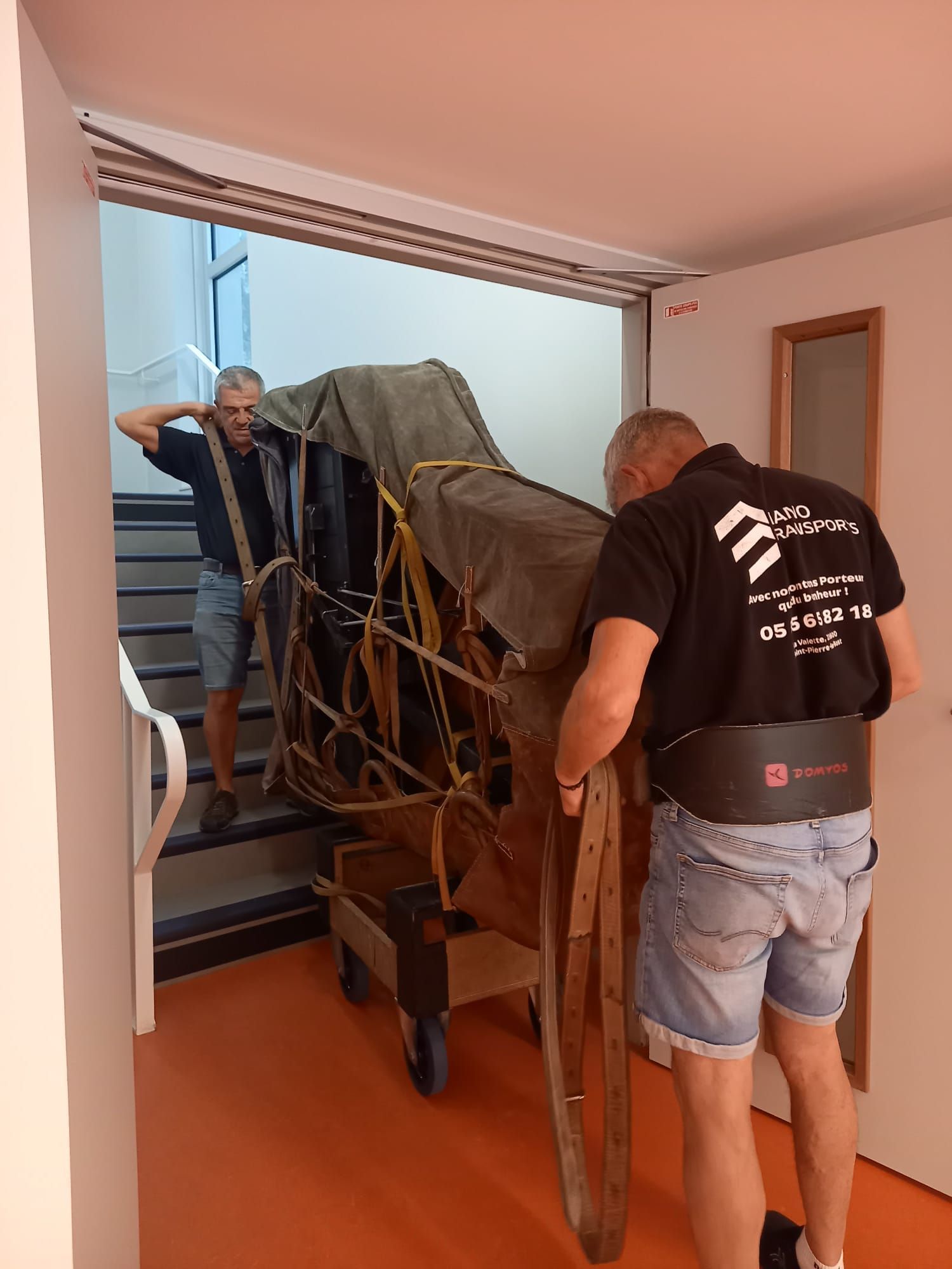Deux hommes manœuvrent une grande charrette en bois à travers une porte dans une cage d'escalier étroite.