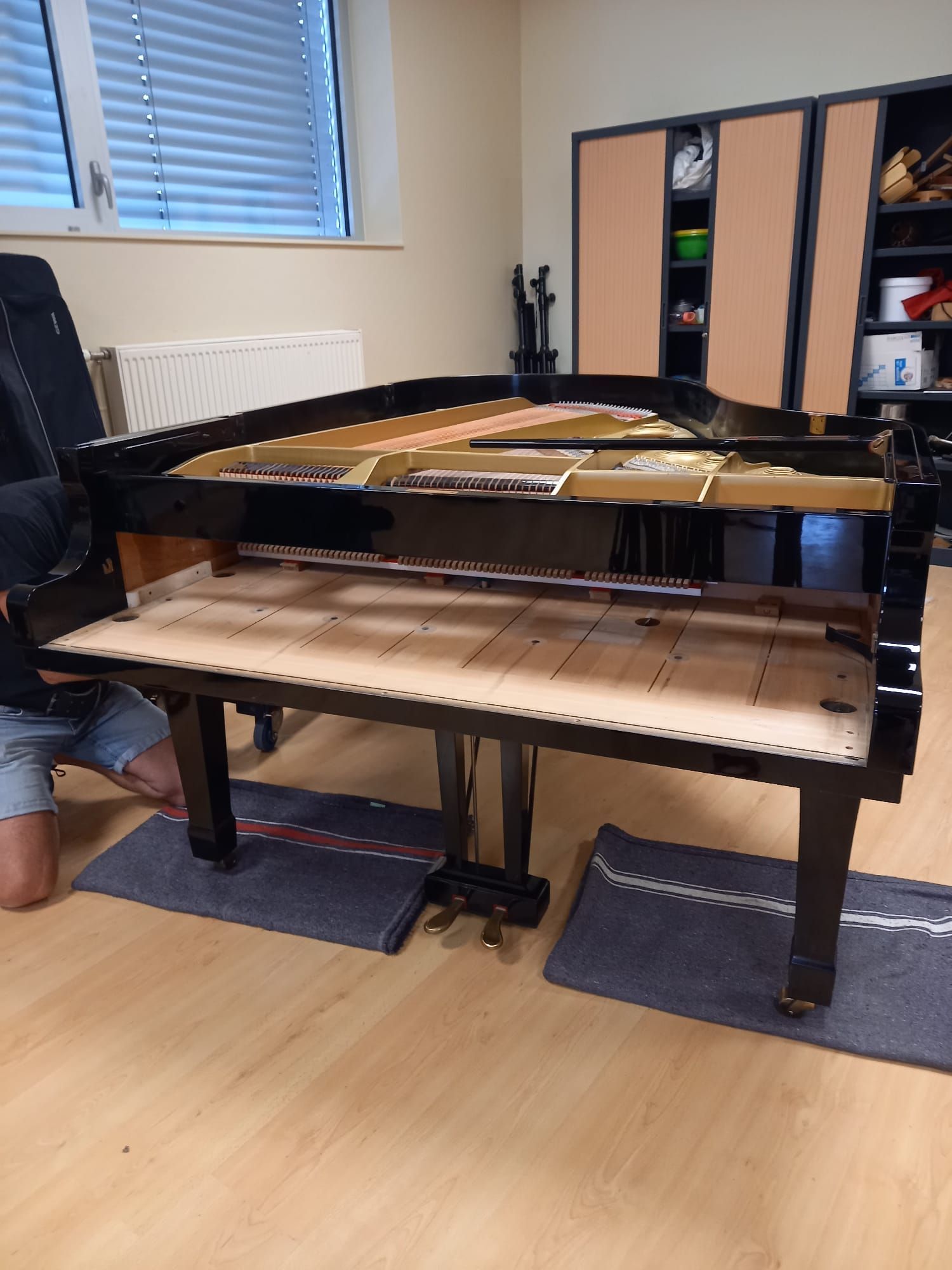 Un piano droit cassé se trouve dans une pièce, les cordes apparentes et le panneau avant détaché gisant au sol.