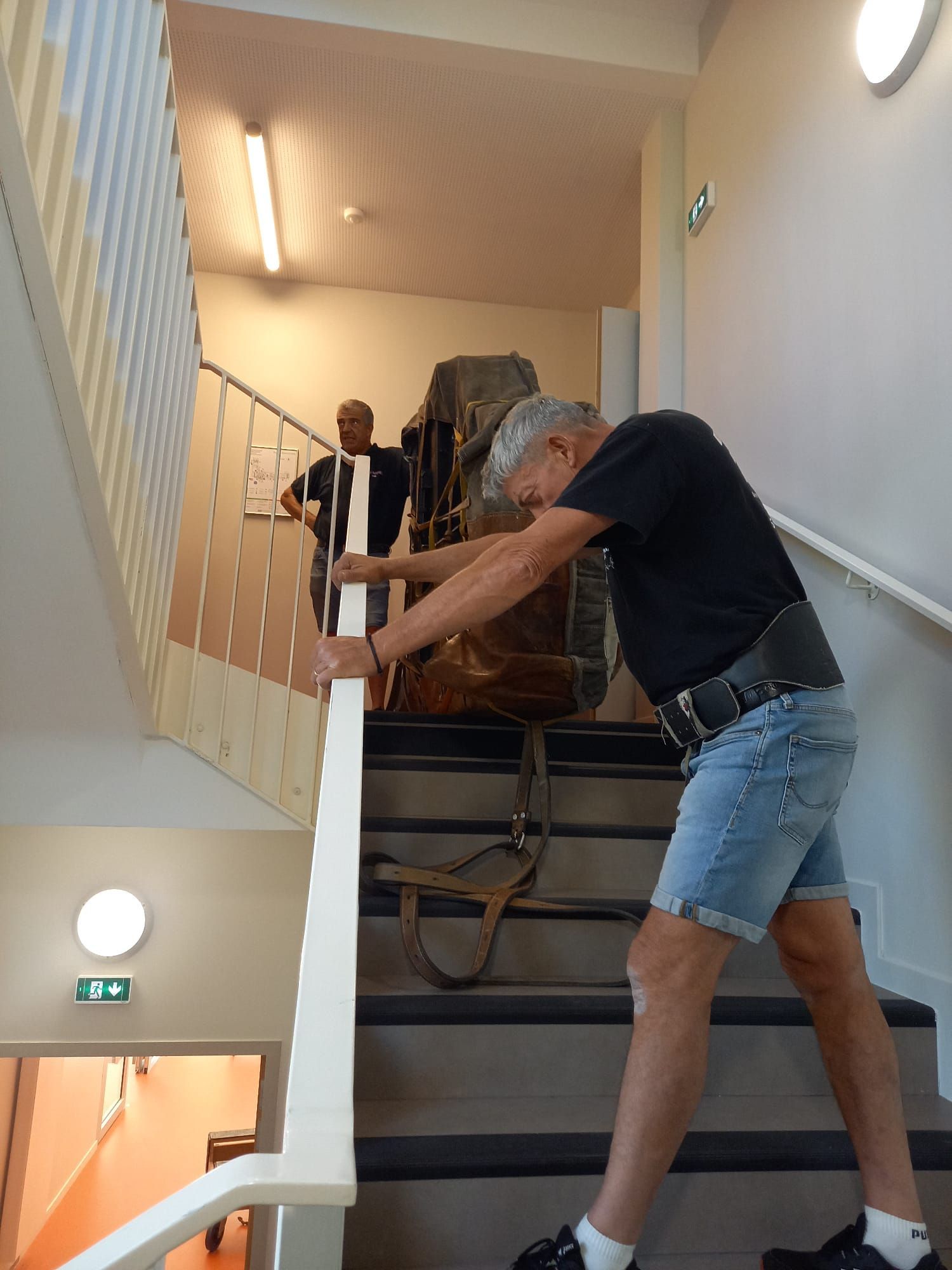 Deux hommes transportant un objet volumineux dans une cage d'escalier à l'intérieur d'un bâtiment.