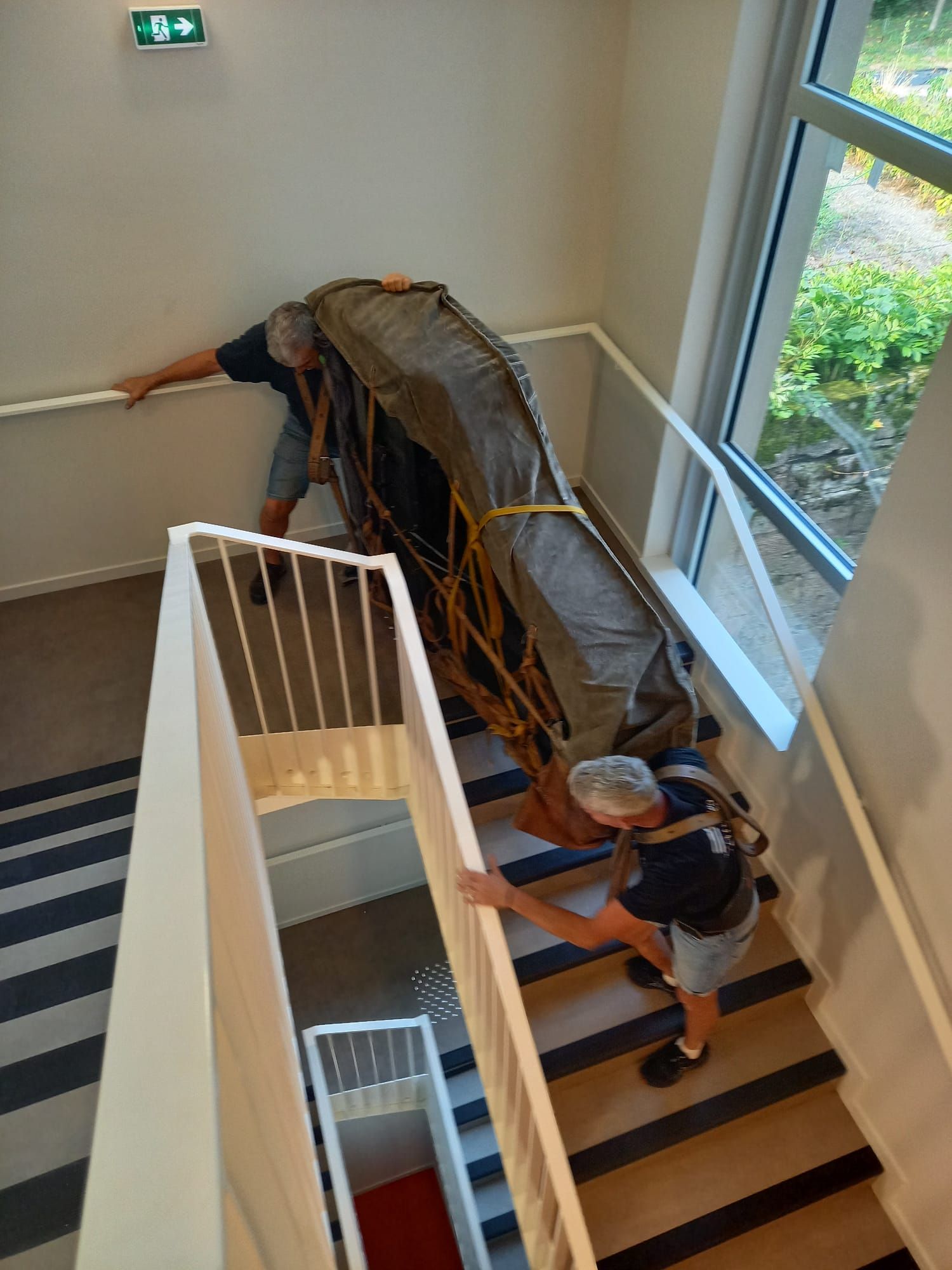 Une personne monte un escalier avec un grand objet drapé, tandis qu'une autre personne le soutient en bas.