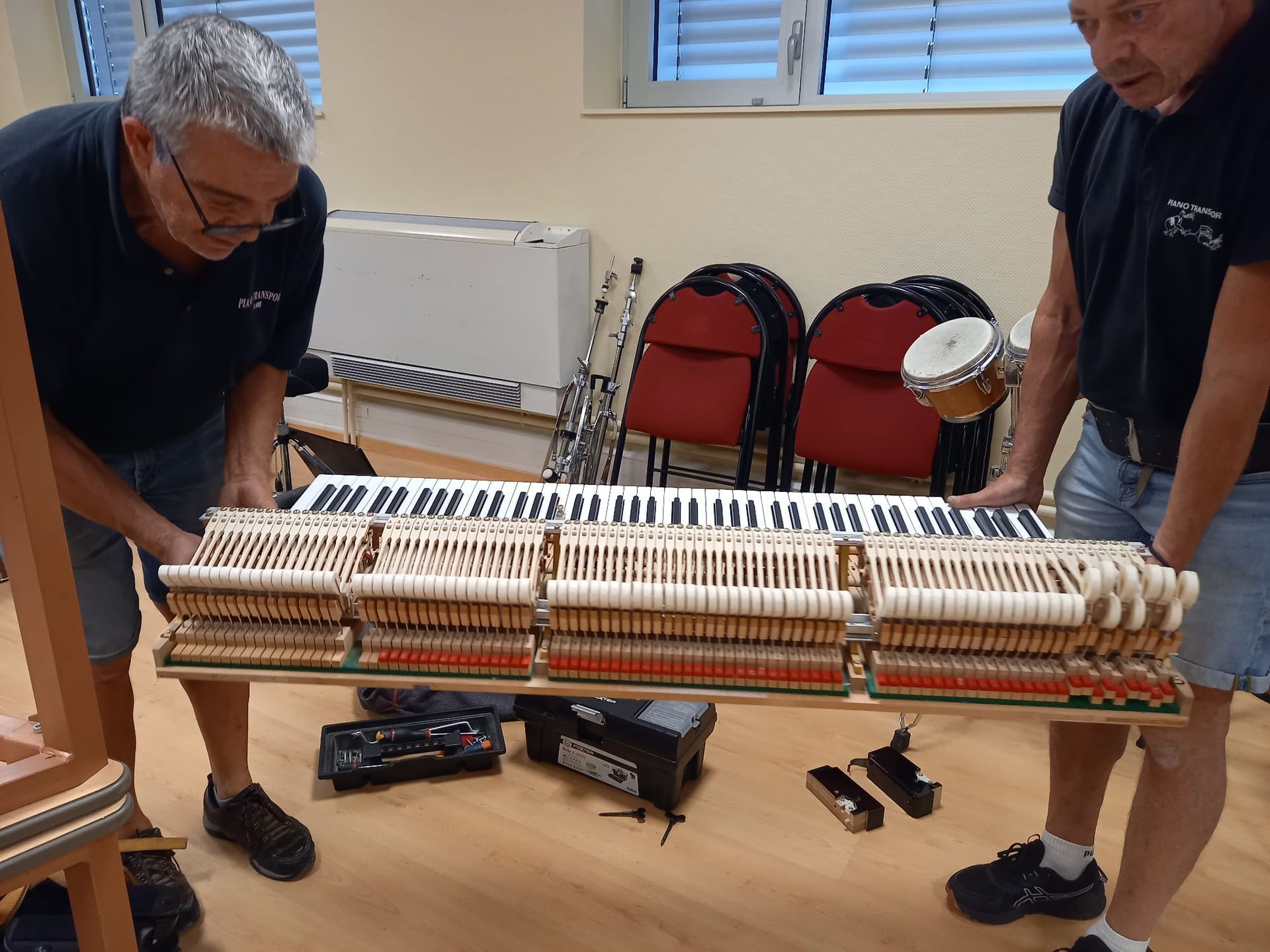 Deux personnes transportant un grand instrument en bois dans une pièce avec des chaises rouges et un radiateur.