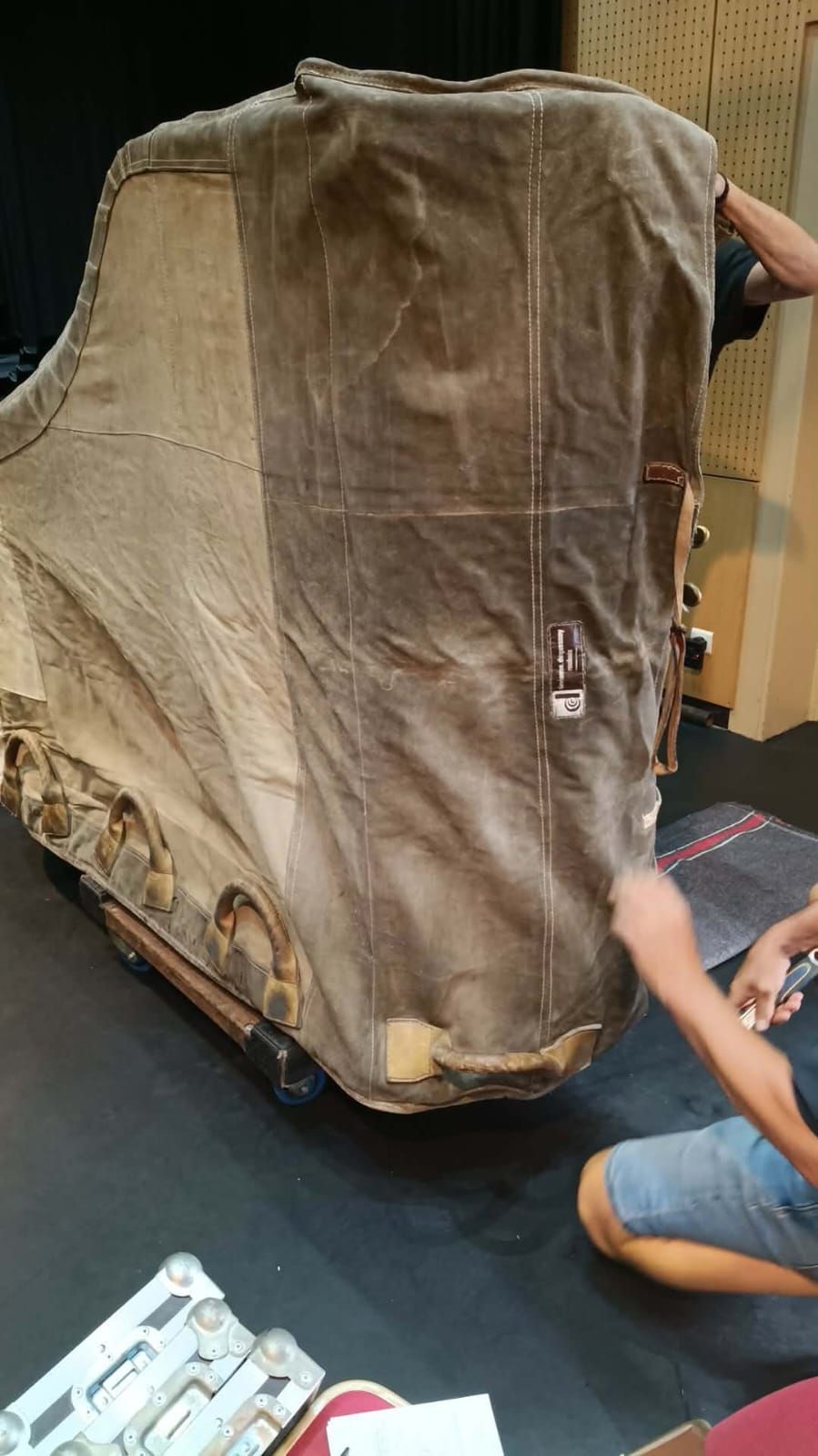Objet volumineux recouvert, monté sur un socle à roulettes, avec une personne agenouillée à côté, dans un atelier intérieur.