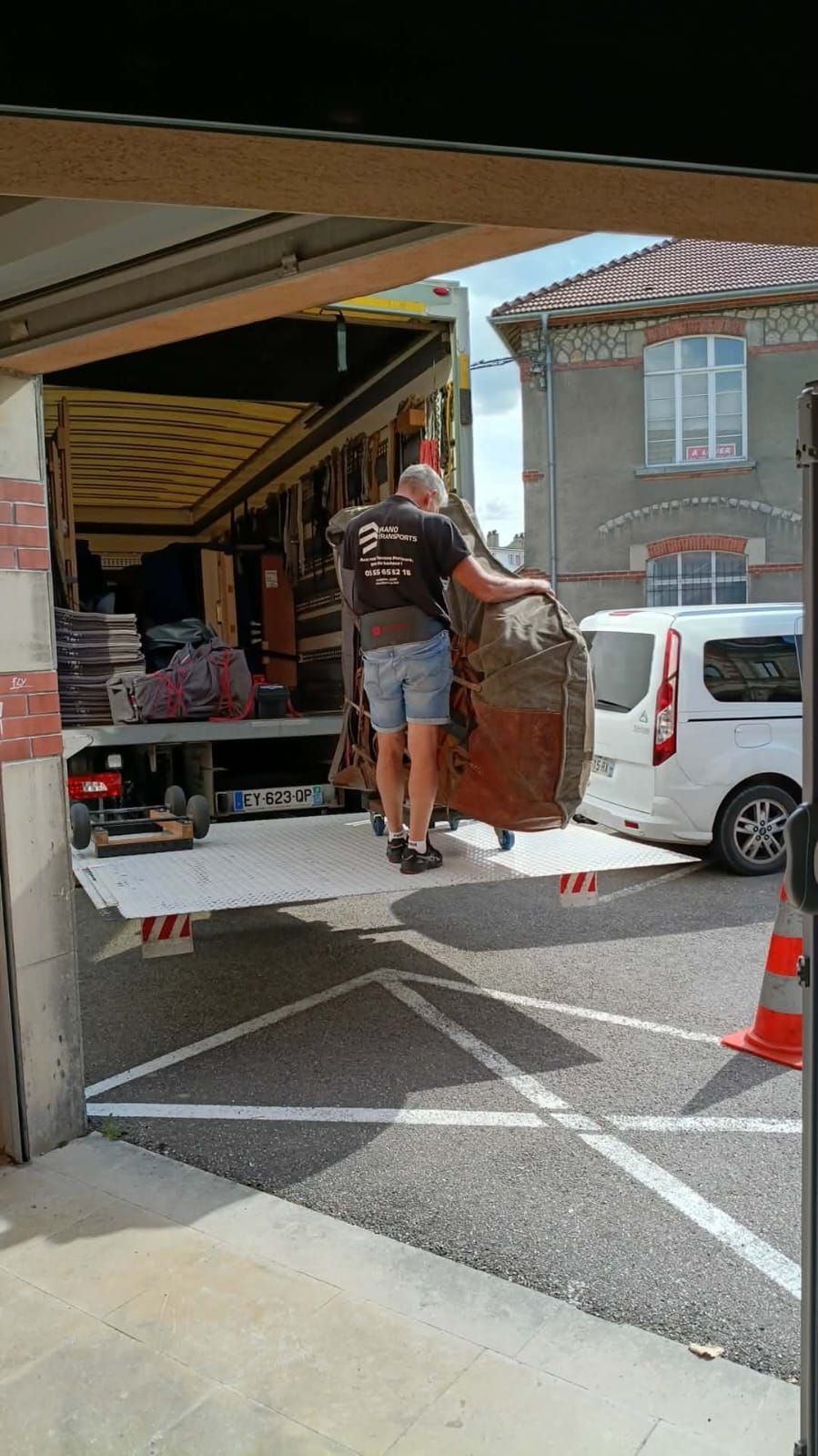 Deux ouvriers déchargent un grand canapé d'un camion dans une allée, à côté d'une camionnette blanche.