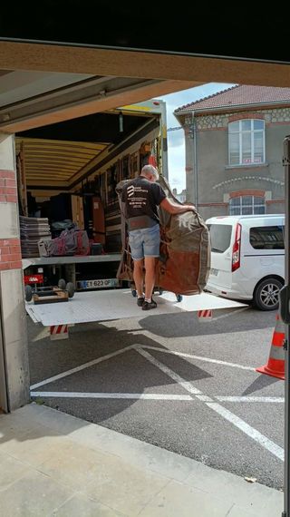 Des déménageurs chargent des meubles dans un camion depuis un garage, une camionnette blanche étant garée à côté.