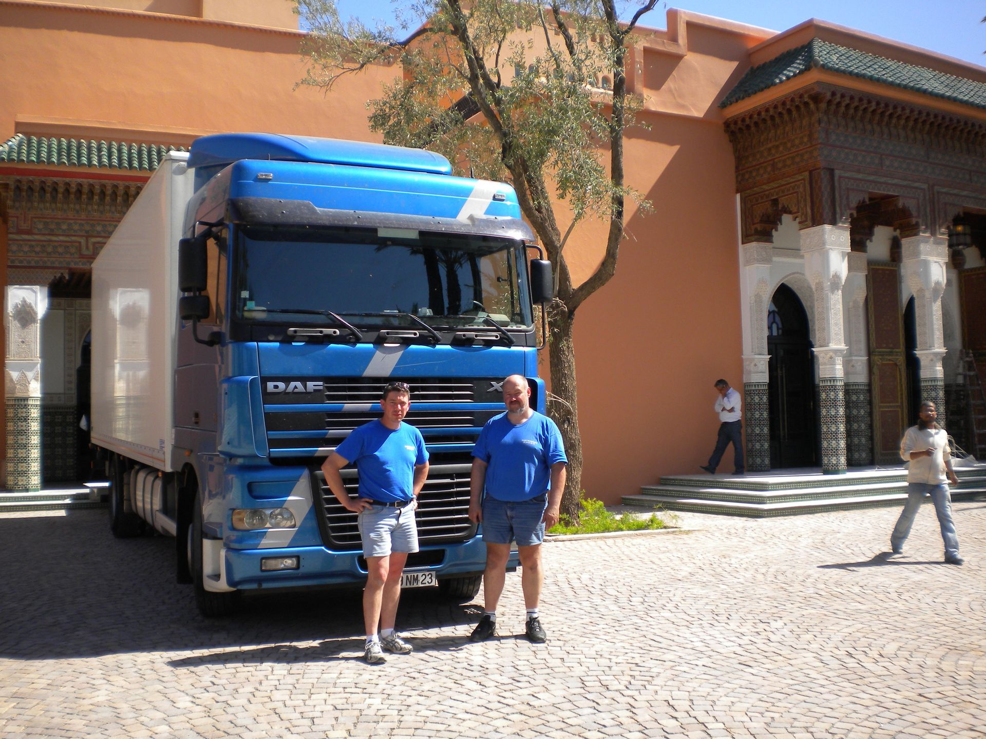 Un camion bleu est garé près d'un bâtiment, deux personnes se tiennent devant.