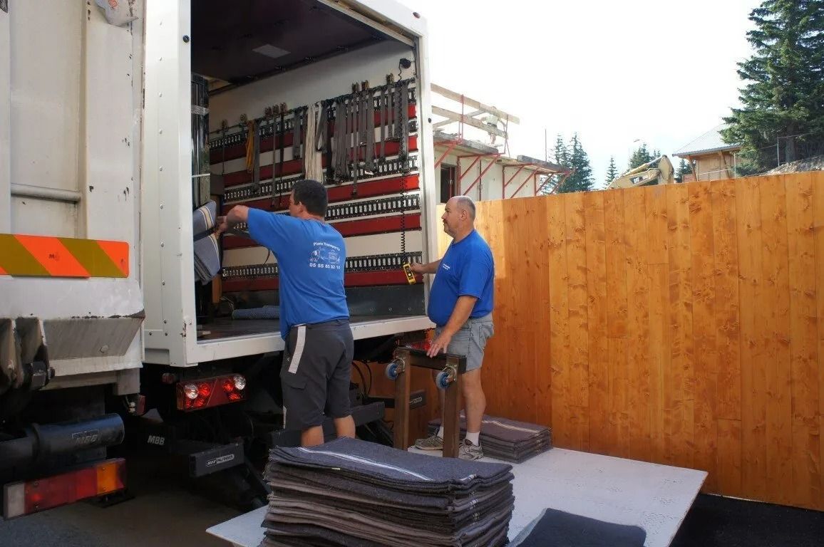 Deux ouvriers chargent des provisions dans un camion blanc près d'une clôture en bois.