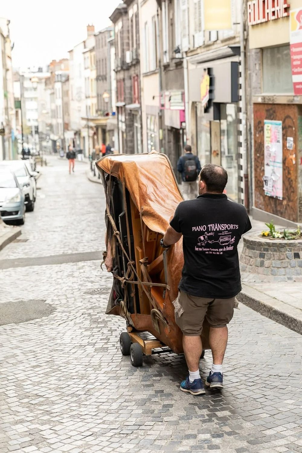 Deux personnes poussent un grand chariot emballé dans une rue pavée étroite bordée de voitures garées et de devantures de magasins.