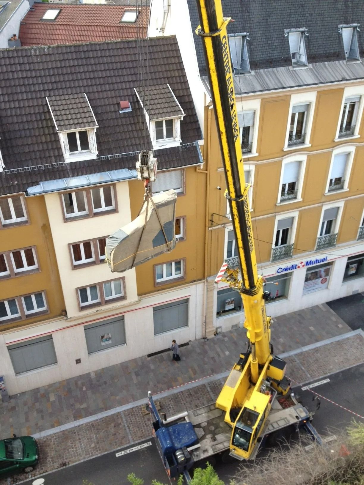Une grue jaune soulève un grand panneau blanc à côté d'un immeuble d'appartements jaune dans une rue de la ville.