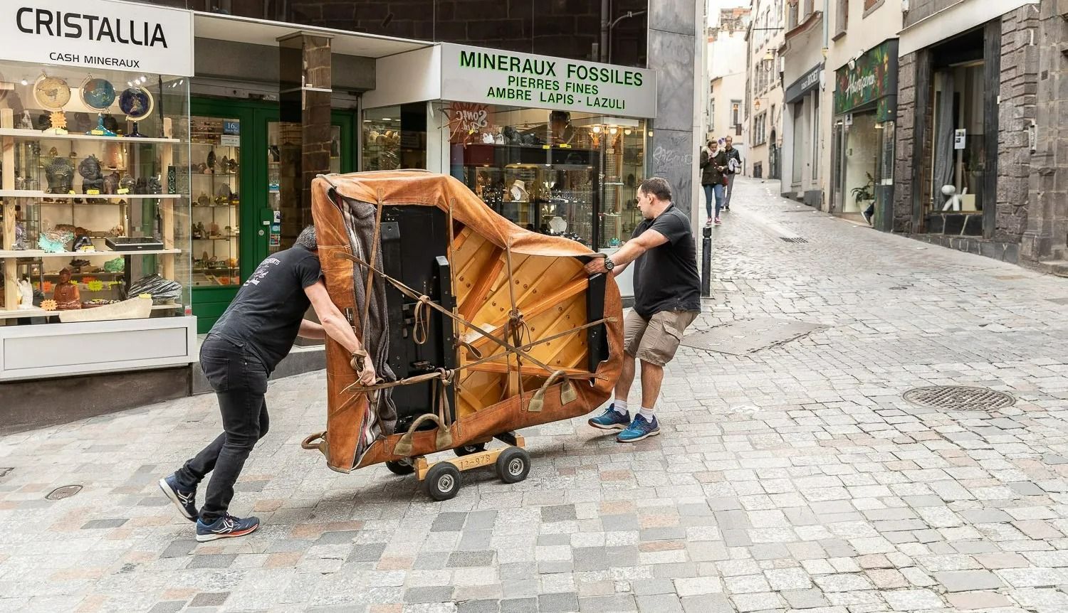 Deux hommes poussent un piano à queue recouvert, monté sur roulettes, dans une rue pavée, le long des devantures de magasins.