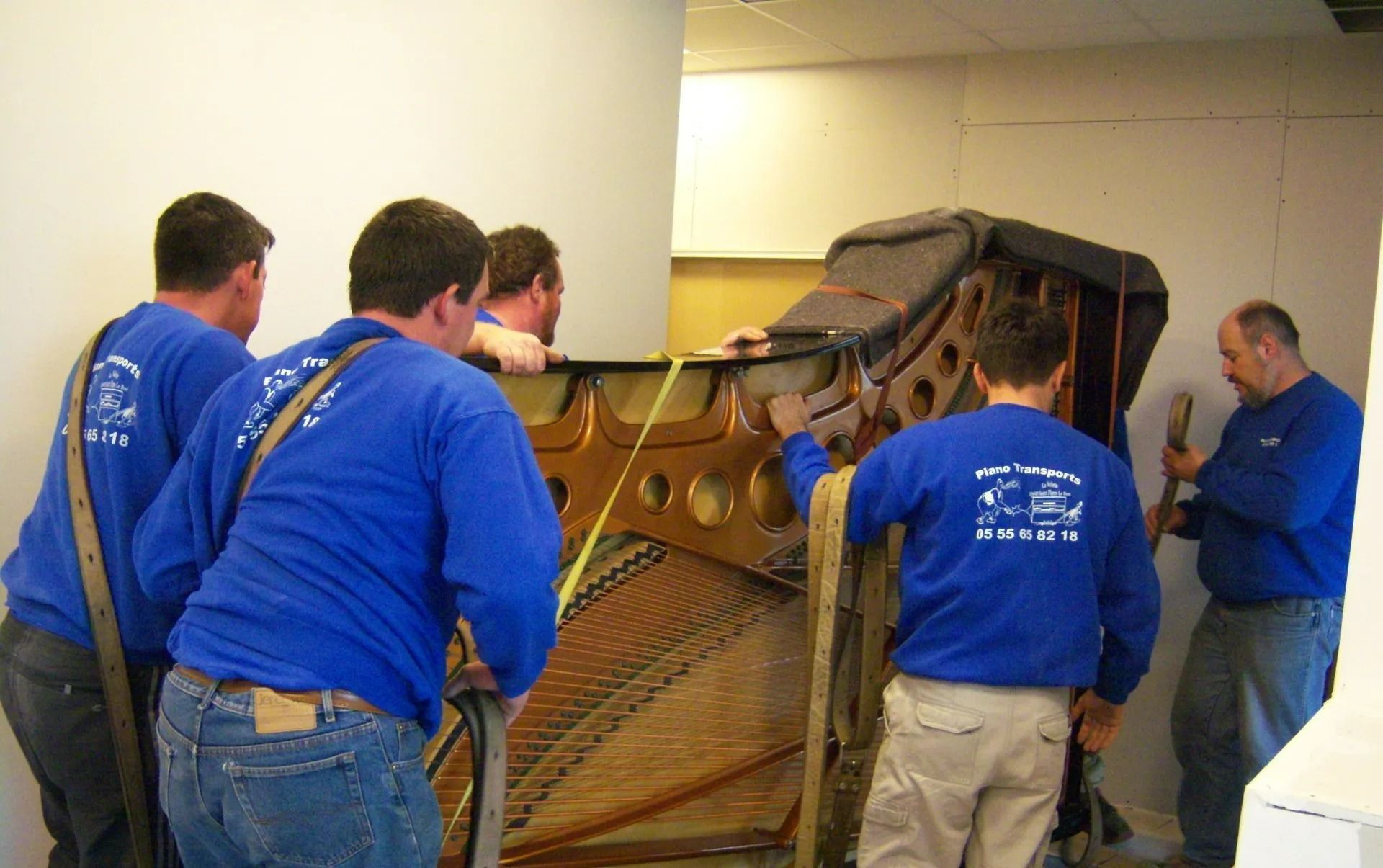 Des ouvriers en chemises bleues déplacent un grand piano droit dans un couloir.