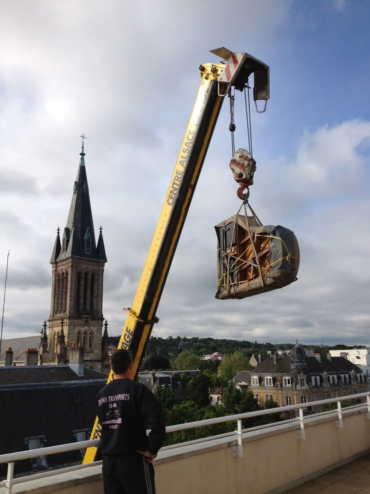 Une grue soulève une cage suspendue au-dessus d'un pont, avec le clocher d'une église en arrière-plan.