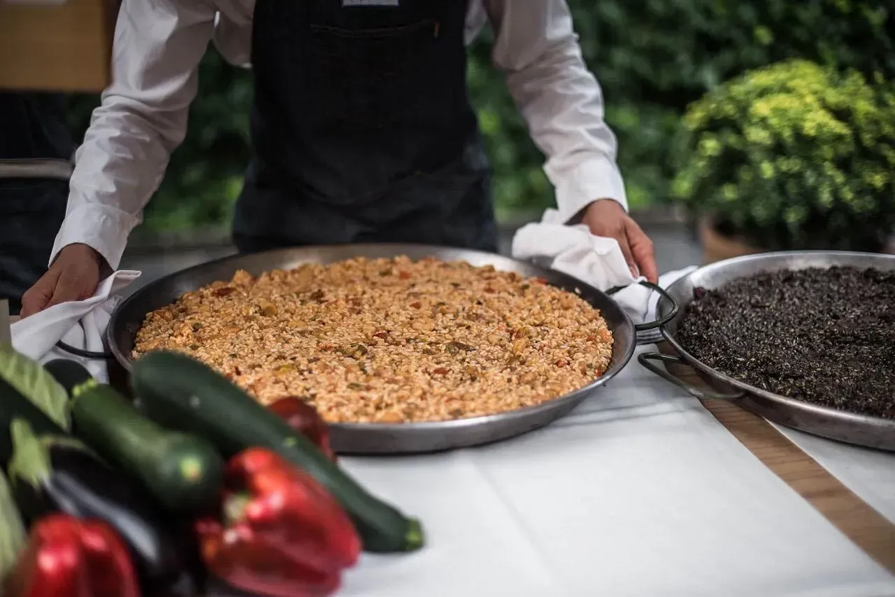 Un hombre está de pie junto a una sartén con comida sobre una mesa.