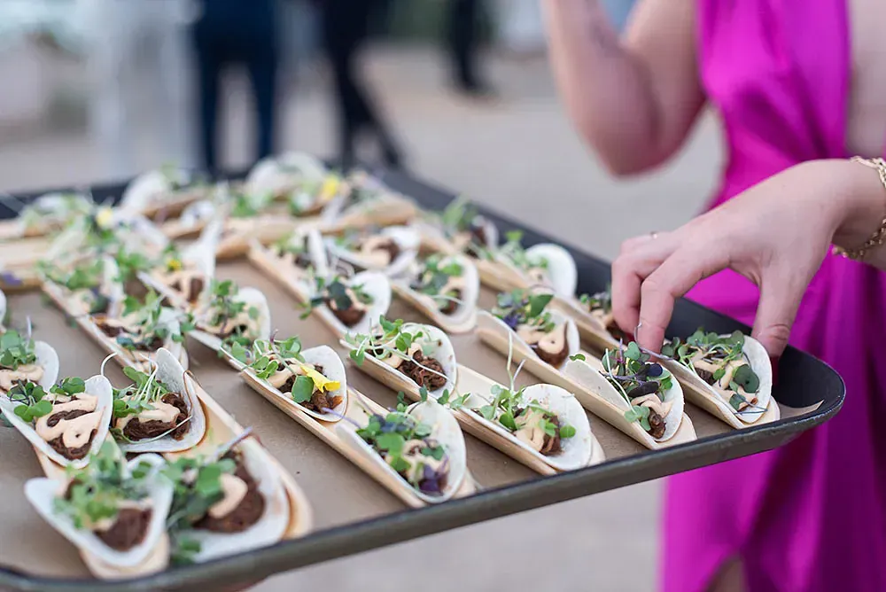 Una mujer con un vestido rosa sostiene una bandeja de tacos.