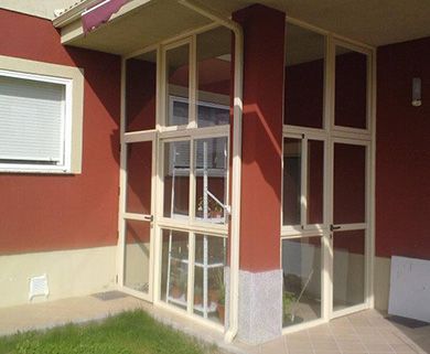 Porche cerrado con paneles de vidrio y marcos de color crema, adosado a un edificio rojo con una ventana blanca.