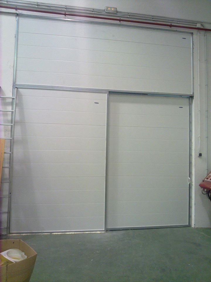 Dos puertas industriales corredizas blancas, una encima de la otra, en un entorno de almacén.