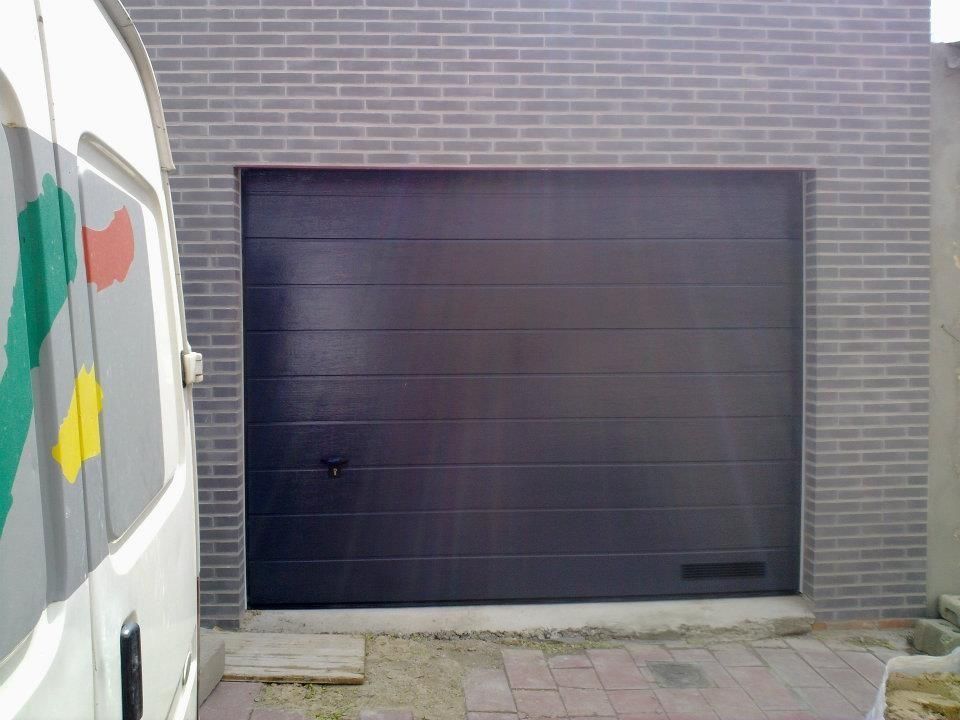 Puerta de garaje negra en una pared de ladrillo gris, al lado de una furgoneta blanca.