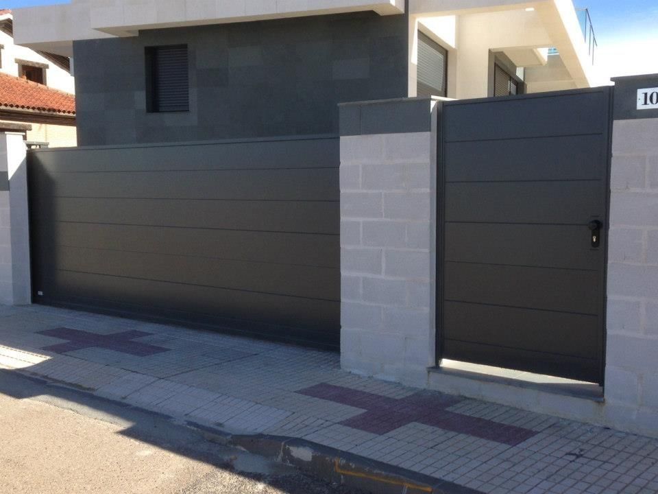 Puerta metálica de lamas horizontales de color gris y puerta peatonal a juego frente a una casa moderna.
