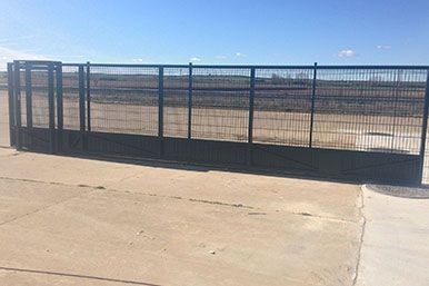 Puerta metálica negra sobre hormigón, abierta, con paneles de malla de alambre, en un entorno exterior bajo un cielo azul.