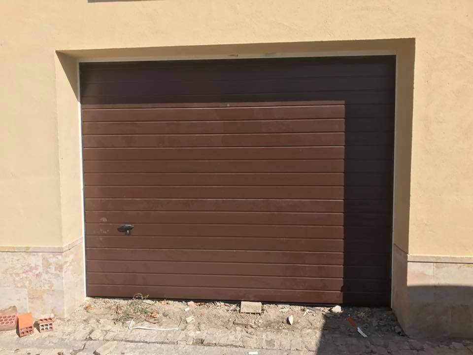Puerta de garaje marrón, parcialmente abierta. El edificio es beige, con cimientos de hormigón y ladrillo.