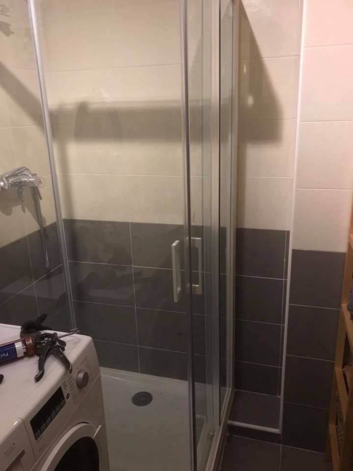 Mampara de ducha en un baño con lavadora visible en la esquina. Azulejos grises y blancos.