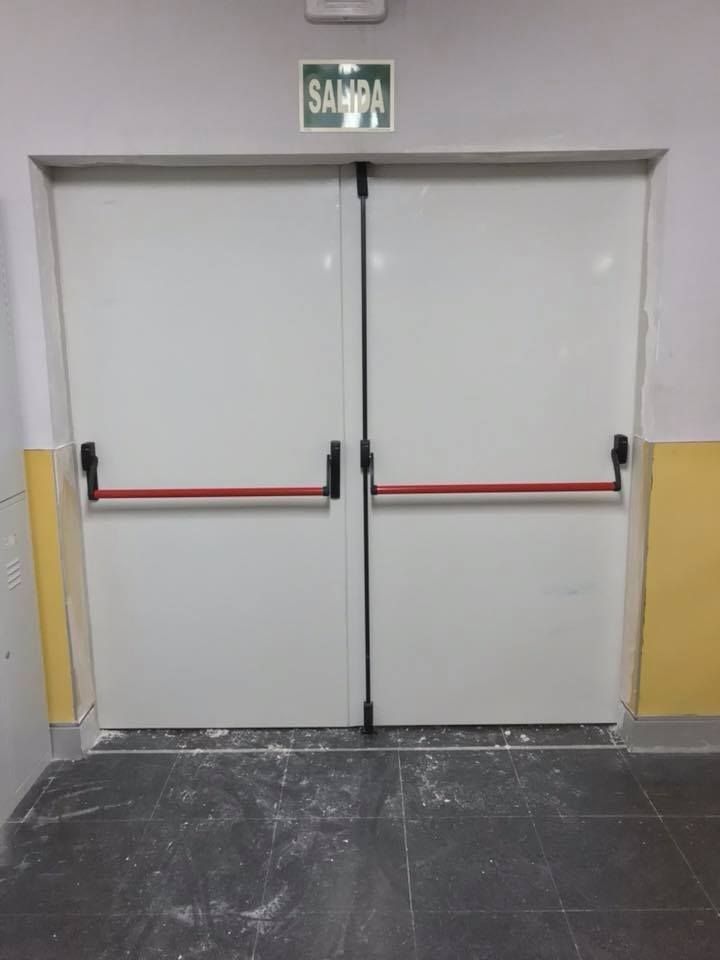 Puertas dobles blancas con barras de empuje rojas, señal de salida encima, en un pasillo.