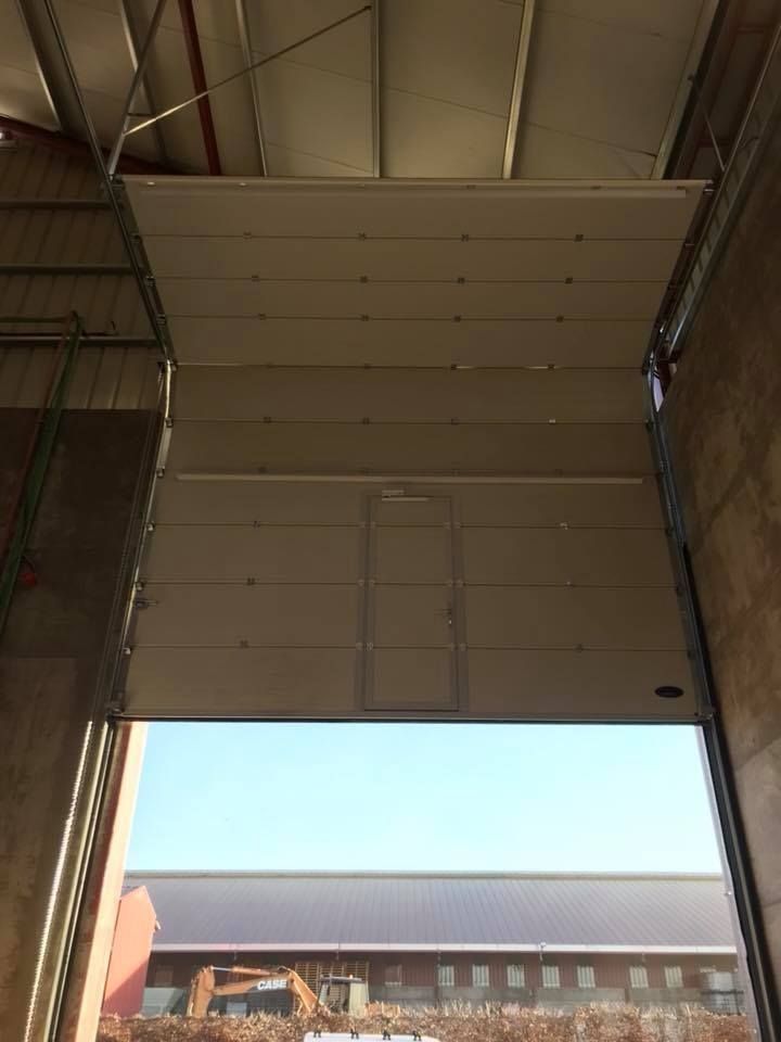 Puerta de almacén grande y abierta con una sección elevada en la parte superior; vista exterior con cielo azul y un edificio a lo lejos.