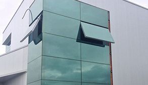Exterior del edificio con paneles de vidrio y dos ventanas con toldo abiertas; reflejo del cielo.