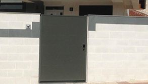 Puerta de metal gris en pared de ladrillo blanco, con pequeño tirador.