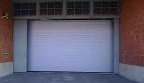 Puerta enrollable industrial de color gris entre paredes de ladrillo, con rejillas de ventilación en la parte superior.