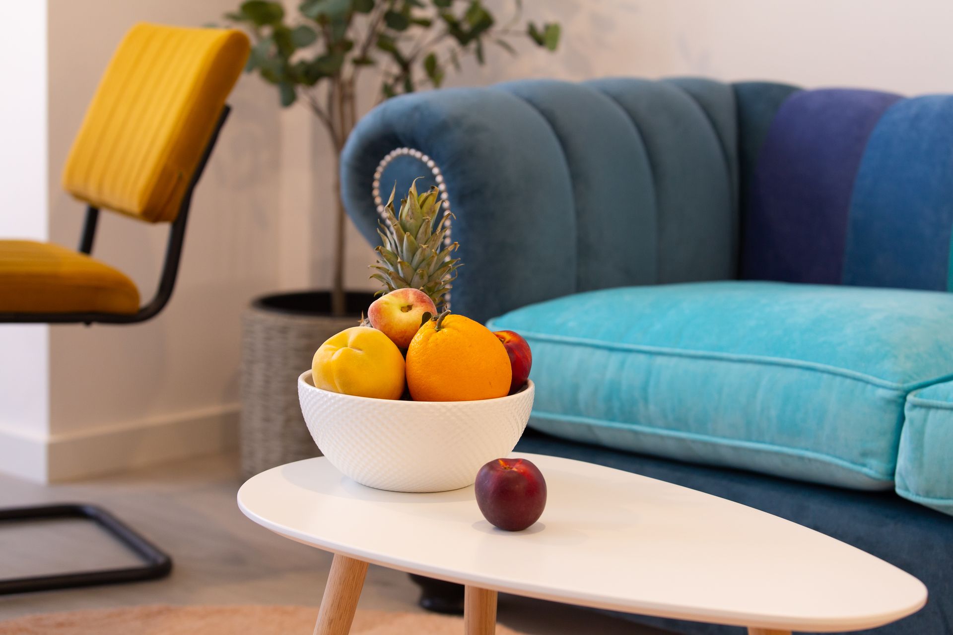 Un bol de fruta reposa sobre una mesa blanca delante de un sofá azul y una silla amarilla.