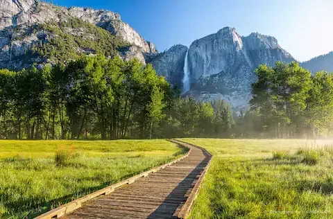 Wooden path through a grassy meadow with trees, leading to a waterfall against a mountain backdrop.